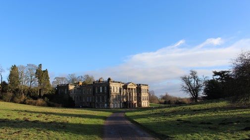 Calke Abbey as seen from the historic drive, surrounded by grassy fields, trees, and framed by blue sky.